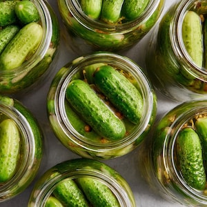 May include: Overhead shot of several glass jars filled with bright green pickles. The pickles are submerged in a clear brine, and the jars are arranged on a light-colored surface. The pickles appear fresh and crisp.