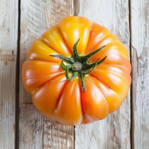 May include: A ripe, heirloom tomato with a ribbed, irregular shape. The tomato is a gradient of orange and yellow hues, with a green stem. The tomato rests on a weathered, light-colored wooden surface.