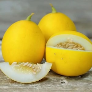 May include: Bright yellow melons, one sliced open to reveal the white flesh and seeds. The other melons are whole, with green stems. The fruit is on a wooden surface.