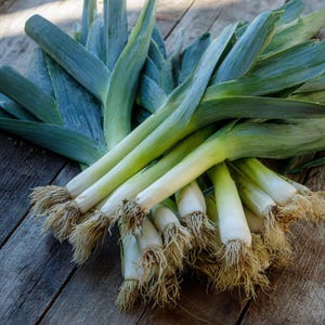 May include: A bundle of fresh leeks with green leaves and white stalks, resting on a weathered wooden surface. The leeks have visible root systems and vary in size, showcasing their natural form.