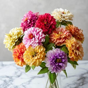 May include: A vibrant bouquet of artificial dahlia flowers in a clear glass vase. The arrangement features blooms in shades of pink, red, orange, yellow, and purple, with green leaves. The flowers are clustered together, creating a full and colorful display.