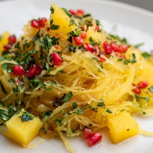 May include: A close-up shot of a plate of spaghetti squash with diced yellow squash, fresh green herbs, and bright red pomegranate seeds. The dish is garnished with chopped parsley and black seeds.