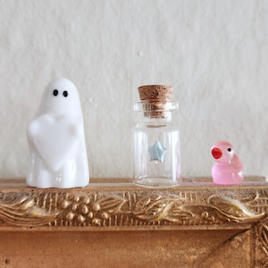 May include: A white ghost figurine holding a heart, a small glass bottle with a cork stopper and a blue star inside, and a pink bird figurine with a red beak are displayed on a decorative gold-colored frame.