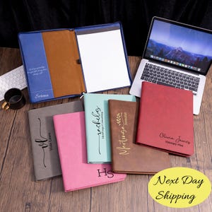 May include: A collection of personalized leather notebooks and portfolios in various colors, including gray, pink, teal, and red. Each notebook features custom text, with a blue portfolio open to reveal lined paper. A laptop and keyboard are also visible.