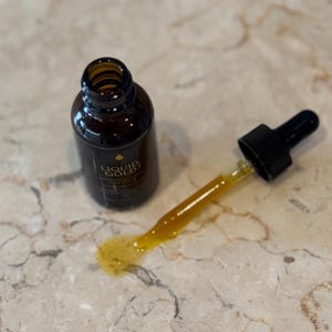 May include: A brown glass bottle with a gold label and the words "LIQUID GOLD" is next to a dropper with a yellow liquid. The dropper is partially out of the bottle and has a black rubber top. A small amount of the yellow liquid is spilled on the surface.