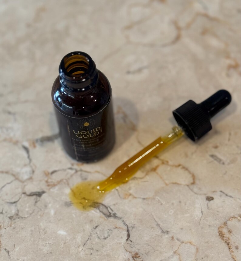 May include: A brown glass bottle with a gold label and the words "LIQUID GOLD" is next to a dropper with a yellow liquid. The dropper is partially out of the bottle and has a black rubber top. A small amount of the yellow liquid is spilled on the surface.