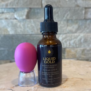 May include: A brown glass bottle of Liquid Gold Radiance-Boosting Face Serum with a black dropper and a pink facial massager. The serum bottle has a black label with gold text. The massager is pink and sits on a clear plastic base.