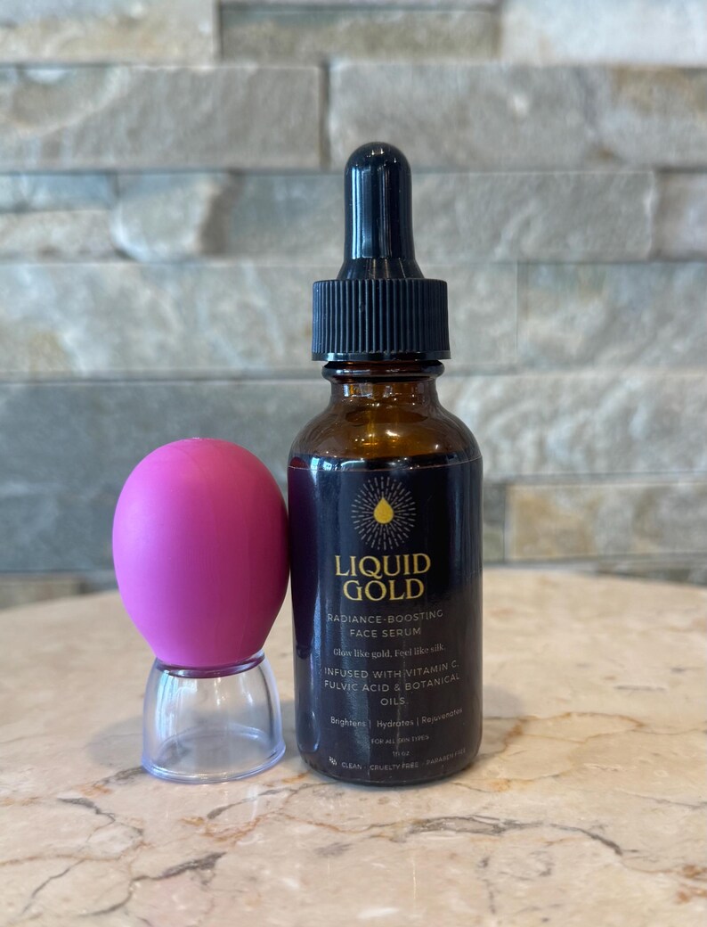 May include: A brown glass bottle of Liquid Gold Radiance-Boosting Face Serum with a black dropper and a pink facial massager. The serum bottle has a black label with gold text. The massager is pink and sits on a clear plastic base.