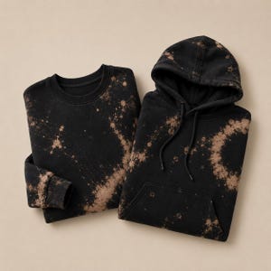 Könnte beinhalten: Ein schwarzes Sweatshirt mit Rundhalsausschnitt und ein passender schwarzer Kapuzenpullover, beide mit einem gebleichten Spritzmuster in Beige. Der Kapuzenpullover verfügt über eine Kängurutasche, eine Kapuze mit Kordelzug und lange Ärmel.