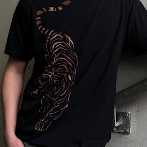 Puede incluir: Camiseta negra con un diseño de tigre color cobre. El tigre está representado en una pose caminando con rayas detalladas. La camiseta tiene mangas cortas y cuello redondo, y se combina con vaqueros azules.