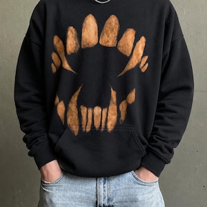 Peut inclure: Sweat à capuche noir avec un grand motif de dents stylisées marron. Le sweat-shirt a une poche avant et une capuche à cordon. Le motif ressemble à des dents de monstre. Le modèle porte un jean bleu.