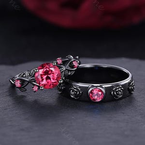 May include: A black engagement ring set featuring a floral design. The engagement ring has a large, round, pink gemstone and smaller pink accent stones. The matching band has rose details and pink gemstones. The rings are set against a dark background.