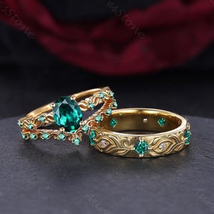 Puede incluir: Dos anillos de oro con piedras preciosas de color verde esmeralda. Un anillo presenta una gran piedra central de esmeralda ovalada, rodeada de esmeraldas más pequeñas. El otro anillo tiene un patrón floral grabado con detalles de esmeralda y pequeños diamantes.