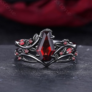 May include: A black ring set with a large, rectangular, deep red gemstone as the centerpiece. The ring is accented with smaller red gemstones and leaf-like designs. The band has a unique, curved shape.