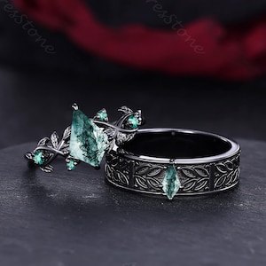 May include: A black wedding ring set featuring a unique design. The engagement ring has a large, irregularly shaped green stone and smaller green accents. The band has a leaf pattern and a teardrop-shaped green stone. The rings are set against a dark background.