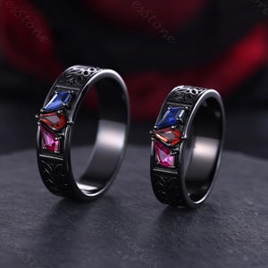 May include: Two black rings with a geometric design. Each ring features three coloured gemstones: blue, red, and pink. The rings have an engraved pattern along the band.