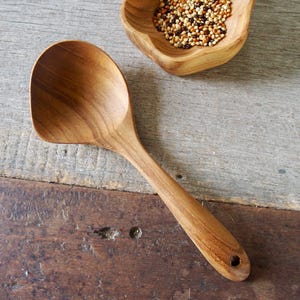 Puede incluir: Una cuchara de cocina de madera con una cabeza ancha y plana y un mango largo. La cuchara es de madera de color marrón claro y tiene un agujero en el mango. Un pequeño cuenco de madera lleno de granos está en el fondo.