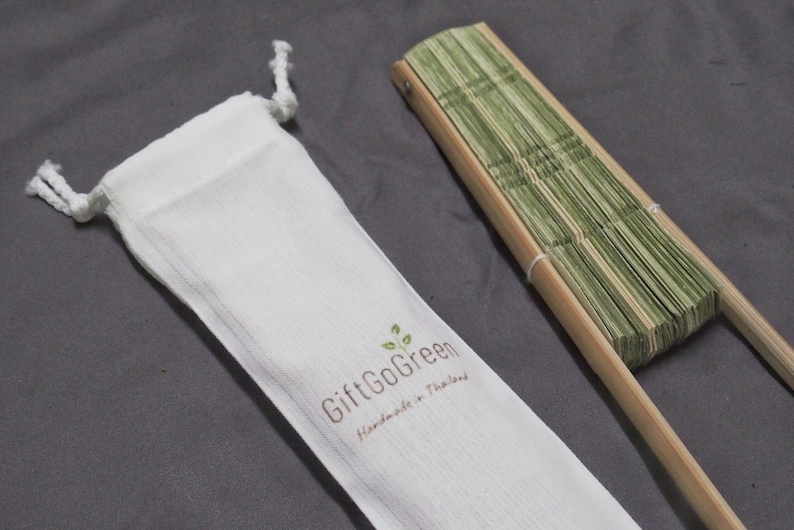 K&ouml;nnte beinhalten: Ein gr&uuml;ner Bambus-Faltf&auml;cher mit einem Holzgriff, kommt mit einem wei&szlig;en Kordelzugbeutel, auf dem "GiftGoGreen Handmade in Thailand" steht.