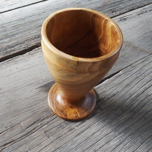 May include: A wooden goblet with a wide, rounded bowl and a flared base. The goblet is made of light brown wood with a natural grain pattern.