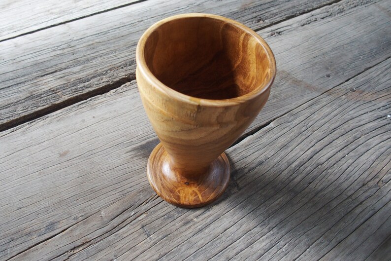 May include: A wooden goblet with a wide, rounded bowl and a flared base. The goblet is made of light brown wood with a natural grain pattern.