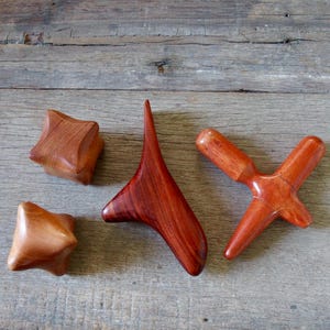 May include: Four wooden massage tools in various shapes and shades of brown. Two are star-shaped, one is a curved triangle, and the last is a cross-shaped tool. The tools are arranged on a weathered wooden surface.