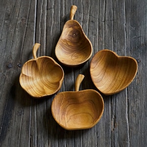 May include: Four wooden bowls shaped like a pear, apple, and heart. The bowls are made of light brown wood and are arranged on a dark brown wooden surface.
