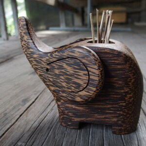 May include: A wooden elephant-shaped toothpick holder with a dark brown finish. The holder has a slot for toothpicks and is made from natural wood.