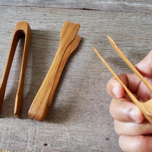 Op de afbeelding: Drie houten tangen, waarvan twee op een verweerd houten oppervlak liggen en één in een hand wordt gehouden. De tangen zijn lichtbruin van kleur en lijken handgemaakt. De tangen zijn ontworpen voor het serveren van voedsel.