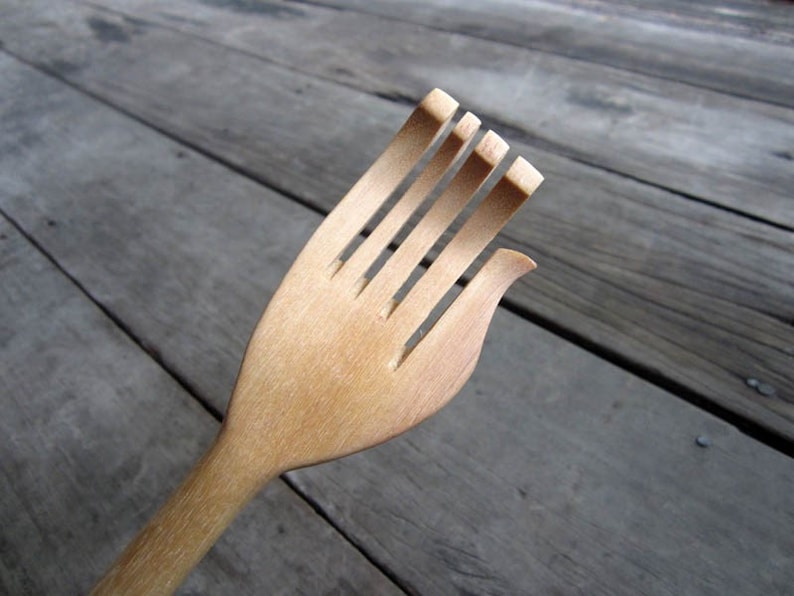 May include: A wooden back scratcher shaped like a hand with five fingers. The scratcher is made of light brown wood and has a long handle.