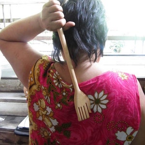 May include: A person is using a wooden back scratcher with a handle and a comb-like end. The scratcher is made of light brown wood. The person is wearing a pink and floral patterned shirt.