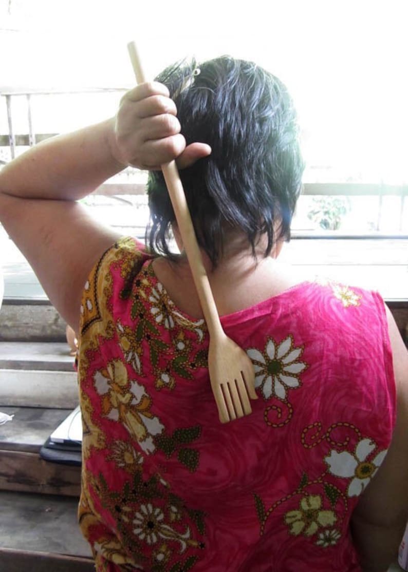 May include: A person is using a wooden back scratcher with a handle and a comb-like end. The scratcher is made of light brown wood. The person is wearing a pink and floral patterned shirt.