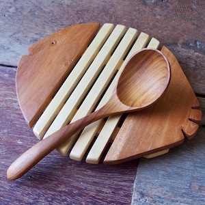 May include: A wooden spoon resting on a circular trivet. The trivet features a combination of light-coloured wooden slats and two darker, reddish-brown wooden side pieces shaped like a fish. The spoon is crafted from a matching reddish-brown wood.