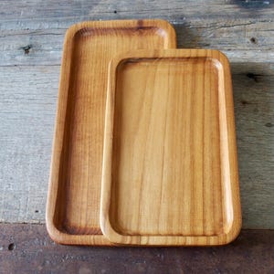 May include: Two rectangular wooden trays with rounded corners. The trays are made of light brown wood and have a smooth, polished finish. The larger tray is behind the smaller one. The trays are on a rustic wooden surface.