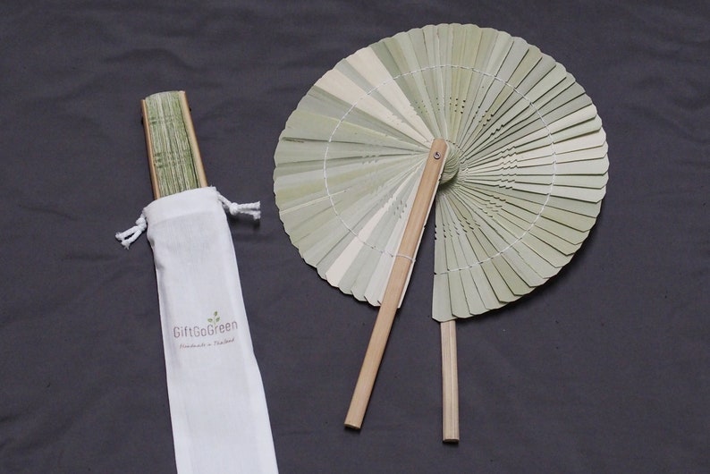 K&ouml;nnte beinhalten: Ein hellgr&uuml;ner Bambus-Handf&auml;cher mit einem Holzgriff. Der F&auml;cher ist zusammengeklappt und wird mit einem wei&szlig;en Kordelbeutel geliefert, auf dem "GiftGoGreen Handmade in Thailand" steht.