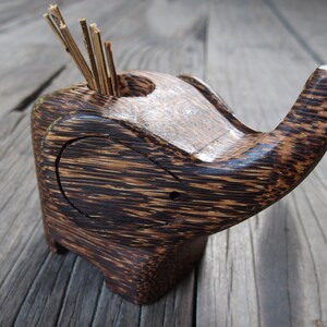 May include: A wooden elephant toothpick holder with a dark brown and black striped finish. The holder has a small opening on the top for toothpicks.