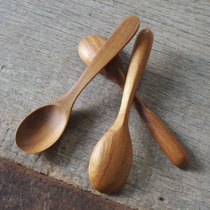 May include: Three wooden spoons with rounded bowls and long handles are arranged on a weathered wooden surface. The spoons are a warm, light brown color, and the wood grain is visible. The spoons are likely for kitchen use.