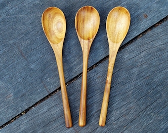 Op de afbeelding: Drie houten lepels op een donkergrijs houten oppervlak. De lepels zijn allemaal even groot en even gevormd, en ze zijn gemaakt van lichtbruin hout.