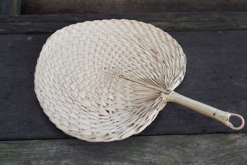 May include: A woven palm leaf fan with a light brown handle. The fan is closed and lying on a wooden surface.
