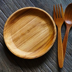 May include: A wooden plate, fork, and spoon set. The plate is round with a shallow bowl. The fork has four tines, and the spoon has a rounded bowl. All pieces are made of light brown wood and rest on a gray wooden surface.