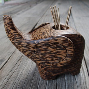 May include: A wooden elephant-shaped toothpick holder with a dark brown and black wood grain pattern. The holder has a small hole in the top for toothpicks, and there are several toothpicks sticking out of the hole.