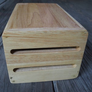 Puede incluir: Una caja rectangular de madera con un acabado claro. La caja tiene dos ranuras horizontales cortadas en la parte delantera. La veta de la madera es visible en la parte superior y los lados. La caja es probablemente para almacenamiento o exhibici&oacute;n.