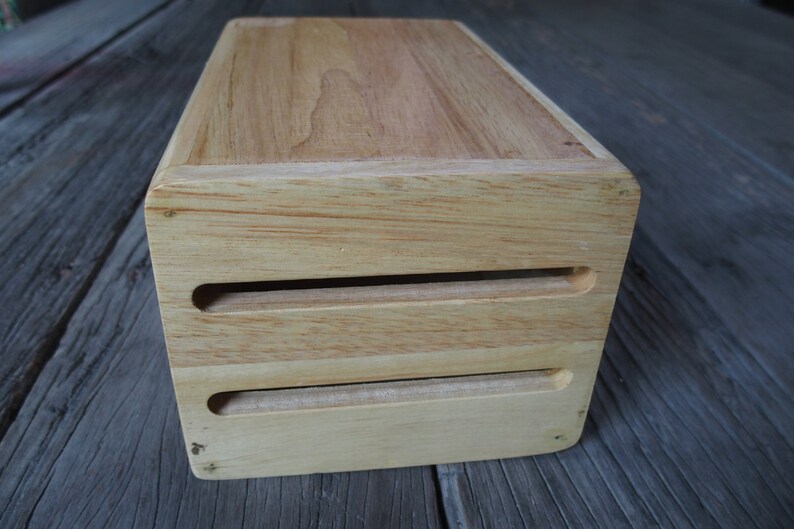 Puede incluir: Una caja rectangular de madera con un acabado claro. La caja tiene dos ranuras horizontales cortadas en la parte delantera. La veta de la madera es visible en la parte superior y los lados. La caja es probablemente para almacenamiento o exhibici&oacute;n.