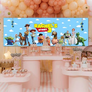 May include: A rectangular party backdrop featuring a blue sky with clouds and a group of animated characters from Toy Story. The text RACHEL'S STORY 3 is printed in yellow and red in the center. The banner is displayed above a dessert table.