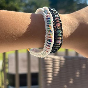 May include: Two bracelets are shown. One is white, the other is black. Both bracelets have a woven design with colorful accents. The bracelets are on a person's arm, with a blurred background of greenery and a sunny sky.