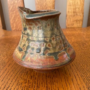 May include: A small, brown, green, and orange ceramic pitcher with a unique, asymmetrical shape. The pitcher has a speckled glaze and a slightly uneven rim.