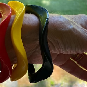 May include: Three wavy bangle bracelets in solid red, yellow, and black colors. Each bangle features a smooth, continuous undulating design. The bracelets are worn together, showcasing their distinct, vibrant colors and uniform, curved, rigid structure.