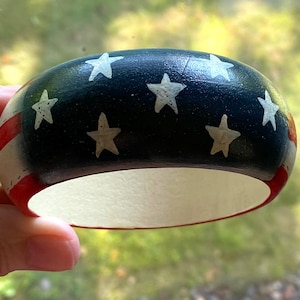 May include: A thick, rounded bangle bracelet featuring a painted design of the American flag. The exterior surface displays a dark blue section with white stars and red and white stripes. The interior of the bangle is a solid, plain off-white color.