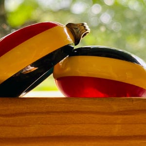 May include: Two smooth, oval-shaped glass ornaments featuring horizontal bands of red, yellow, and black. One ornament rests horizontally while the other leans against it at an angle, both displaying a glossy, reflective surface finish.