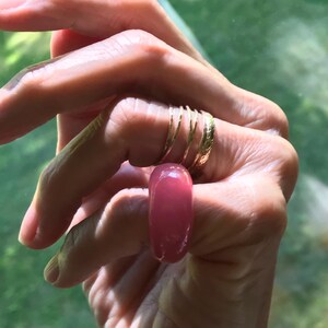 May include: A set of four gold-toned rings worn on the fingers, including three thin, smooth bands and one wider band with a textured surface, paired with a single, smooth, opaque pink resin ring worn on the adjacent finger.