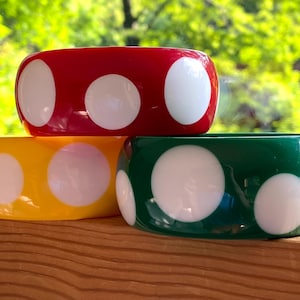 May include: Three thick bangle bracelets with a glossy finish, each featuring a solid color base of red, yellow, or green with large, circular white polka dot patterns distributed evenly across the exterior surface of the rounded bands.